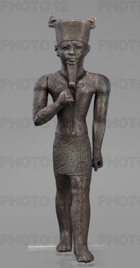 Figurine of the god Amun, Amun mythology, Egyptian deities, male figurines, Egyptian art, Once holding a khepesh, ca. 1069–ca. 747 BC; Third Intermediate Period, Egypt, sculpture, height 19.6 cm, width 6.2 cm, A standing male figure sculpture with a tall flat-topped headdress and a stylized beard held by one hand against the chest, rendered in a rough textured dark material that appears brownish-black with some lighter brown highlights; the figure wears a short patterned skirt with carved linear and diagonal motifs and a collar-like necklace, has one arm bent across the chest and the other hanging by the side, and shows detailed facial features and ears; the base and background are neutral gray, and the surface shows patches of lighter brown and subtle metallic sheen.
