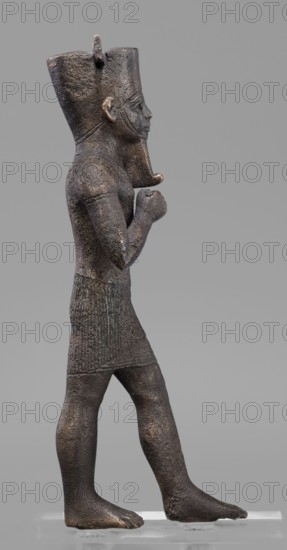 Figurine of the god Amun, Amun mythology, Egyptian deities, male figurines, Egyptian art, Once holding a khepesh, ca. 1069–ca. 747 BC; Third Intermediate Period, Egypt, sculpture, height 19.6 cm, width 6.2 cm, A dark brown metal statue of a standing figure shown in profile, facing right, with a tall rounded headdress and a stylized beard; the figure has a textured surface with areas of lighter brown and subtle golden highlights, visible facial features and a prominent ear, both arms bent with closed fists held near the chest, a short textured skirt, and bare legs and feet planted on a pale rectangular base against a neutral gray background.