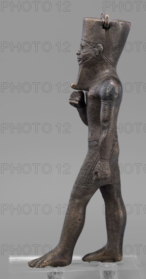 Figurine of the god Amun; maker, Amun mythology; Egyptian deity male figurine; once held a khepesh; ca. 1069–ca. 747 BC, Third Intermediate Period, Egypt; sculpture; height 19.6 cm, width 6.2 cm., A dark brownish bronze statue of a standing male figure shown in profile, facing left, wearing a tall headdress and a short textured skirt; the figure has a stylized beard and a pronounced jawline, a closed mouth, and a visible ear, with one arm bent and fist near the chest and the other arm hanging down with a clenched hand; the surface shows a rough, pitted texture with areas of lighter brown and subtle greenish or blackish patina; the figure stands barefoot on a clear rectangular support against a plain light gray background.