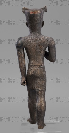 Figurine of the god Amun, Amun mythology, Egyptian deities, male figurine, Egyptian art, once holding a khepesh, ca. 1069–ca. 747 BC; Third Intermediate Period, Egypt, sculpture, height 19.6 cm, width 6.2 cm, A bronze figurine viewed from the back showing a standing human figure with a tall flat-topped headpiece, short hair or a smooth scalp, and stylized ears; the surface is textured and pitted with a dark brown to black patina and areas of lighter brown highlights, and there is a carved collar pattern across the upper back and a patterned skirt around the hips with vertical ribbing; the left arm is bent at the elbow and the right arm hangs down with a closed hand holding a short hollow cylindrical object, the legs are straight with visible calf musculature, and the figure stands on a clear rectangular base against a uniform gray background.