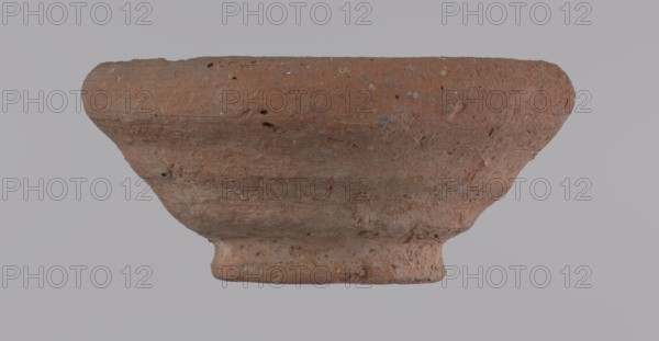 Bowl, Egyptian pottery, clay vessel, bowl, 332 BC–395 AD Ptolemaic–Roman Period, from Edfu; vessel; height 5 cm; rim diameter 11 cm; bottom diameter 5.7 cm., A worn, shallow bowl-like object with a rough, porous surface in shades of reddish-brown and muted orange, showing patches of darker brown and small areas of gray; the form has a flared rim, a gently curved body, and a narrower foot with uneven texture, with visible pits and small holes across its surface against a pale gray background.