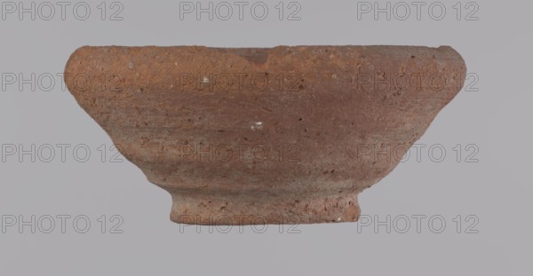 Bowl, Egyptian pottery bowl of clay; 332 BC–395 AD Ptolemaic–Roman Period; from Edfu; vessel; height 5 cm; rim diameter 11 cm; bottom diameter 5.7 cm., A single earthenware bowl is centered against a plain light gray background, showing a rough textured surface with visible pits and small inclusions; the bowl has a wide flared rim that narrows toward a short, slightly raised base, and its surface displays a range of reddish-brown and terracotta tones with areas of darker brown and small pale specks.