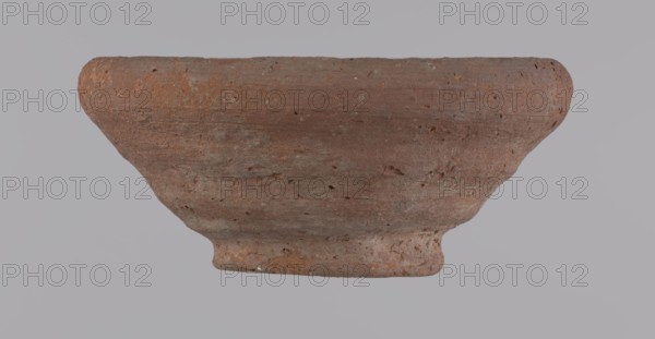 Bowl, Egyptian pottery vessel, clay bowl; 332 BC–395 AD Ptolemaic–Roman Period; from Edfu; height 5 cm, rim diameter 11 cm, bottom diameter 5.7 cm., A textured reddish-brown clay bowl with a rounded rim and a short foot, showing surface pitting and irregularities, set against a pale gray background; colors visible include reddish-brown, gray, and subtle darker brown spots.