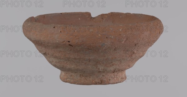 Bowl, unknown creator; Egyptian pottery vessel made of clay; bowl; Ptolemaic Period–Roman Period 332 BC–395 AD; from Edfu; height 5 cm; rim diameter 11 cm; bottom diameter 5.7 cm., A weathered clay bowl with a rough, pitted surface sits against a plain light gray background; the bowl is mostly reddish-brown with patches of darker brown and small flecks of lighter beige, showing irregular texture and small holes, a slightly flared rim with uneven edges, a rounded body that tapers to a short, slightly raised foot, and visible surface variations and chips.