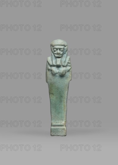 Ushabti, Mummy-shaped funerary figurine shabti, standing on a base, back supported by a pillar/pilaster. Arms crossed on the chest, right folded over left; both hands holding hoes. A bag is on the back, slung over the left shoulder on a single string. Wearing a three-part smooth wig with the back strand longer than the front strands; long, straight, wide beard. Large eyes, wide nose, mouth set in a grimace, very large ears. No inscription. Late Period, Egypt, 664–332 BC. Height 7.7 cm; width 2.1 cm., A small carved figurine stands upright against a plain gray background, showing a stylized human form with a headpiece and a long, tapered body resting on a short rectangular base; the surface is predominantly a bluish-green with areas of muted green and pale gray, and the figure's facial features and folded arms are roughly modeled with subtle texture and weathered patina.