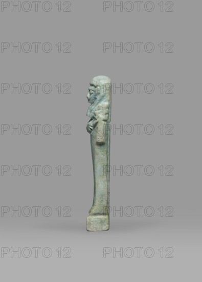 Ushabti, Mummy-shaped funerary figurine shabti standing on a base, back supported by a pillar/pilaster. Arms crossed on the chest, right folded over left; both hands holding hoes. A bag is on the back, slung over the left shoulder on a single string. Wearing a three-part smooth wig, the back strand longer than the front strands. Long, straight, wide beard; large eyes, wide nose, mouth set in a grimace, very large ears. No inscription. Late Period, Egypt, 664–332 BC. Height 7.7 cm, width 2.1 cm., A tall, narrow statuette shown in profile against a plain background, rendered in greenish-blue tones with areas of paler and darker green; the figure stands on a small rectangular base and has a long, slender body with a straight posture; the head is rounded with visible facial features in low relief and a headdress or hair that falls down the back; the arms are bent and held close to the torso with hands resting near the abdomen; the surface shows a weathered, slightly rough texture with subt...