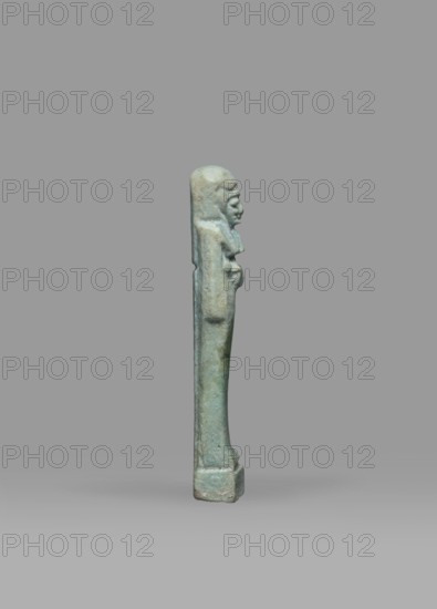 Ushabti, Mummy-shaped funerary figurine shabti, standing on a base, back supported by a pillar/pilaster. Arms crossed on the chest, the right folded over the left; both hands holding hoes. A bag is on the back, slung over the left shoulder on a single string. Wearing a three-part smooth wig with the back strand longer than the front strands; long, straight, wide beard. Large eyes, wide nose, mouth set in a grimace, very large ears. No inscription. Late Period, 664–332 BC, Egypt. Height 7.7 cm; width 2.1 cm., A small upright statuette of a human figure shown in profile against a plain gray background; the figure is narrow and elongated with a rounded head, pronounced facial features, and arms held close to the torso, standing on a rectangular base. The surface shows a weathered, mottled greenish-blue color with areas of lighter and darker green and hints of brownish-gray patina. The background is a uniform medium gray with a slightly lighter gray ground beneath the base.