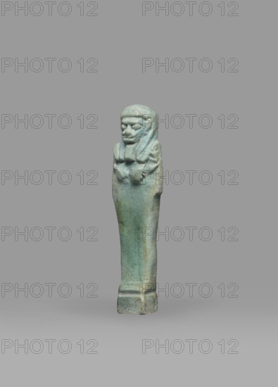 Ushabti, Mummy-shaped funerary figurine shabti, standing on a base, back supported by a pillar/pilaster. Arms crossed on the chest, right folded over left; both hands holding hoes. On the back is a bag slung over the left shoulder on a single string. Wearing a three-part smooth wig with the back strand longer than the front strands. Long, straight, wide beard; large eyes, wide nose, mouth set in a grimace, very large ears. No inscription. Late Period, Egypt, 664–332 BC. Height 7.7 cm; width 2.1 cm., A small figurine standing upright on a flat base, depicting a mummiform figure with a stylized head and headdress, arms crossed over the chest, and a long tapered body; the surface shows a weathered, slightly rough texture with visible wear and small chips, and the colors visible are shades of turquoise, green, and pale blue with areas of darker green and muted beige.