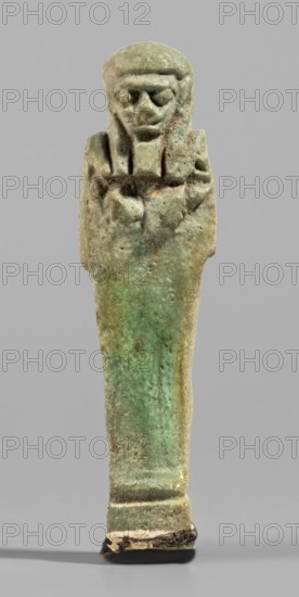 Ushabti, Mummy-shaped funerary figurine standing on a base, back supported by a pillar/pilaster. Arms crossed on the chest, right folded over left; both hands holding hoes. Bag on the back slung over the left shoulder on a single string. Three-part smooth wig back strand longer than front strands. Long, straight, wide beard. Large eyes, wide nose, mouth set in a grimace, very large ears. No inscription. Late Period, 664–332 BC, Egypt. Shabti. Height 7.7 cm; width 2.1 cm., A small carved figure stands upright against a plain gray background, showing a stylized human form with a headdress and crossed arms; the surface has weathered texture and a patina ranging from pale green to turquoise with areas of brown and beige, and darker spots and rougher brownish edges near the base.