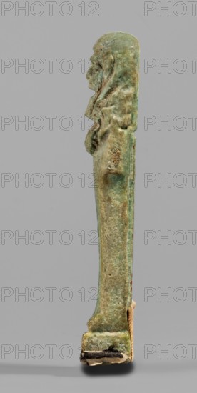 Ushabti, Mummy-shaped funerary figurine, standing on a base with back supported by a pillar/pilaster. Arms crossed on the chest, right folded over left; both hands holding hoes. A bag is slung over the left shoulder on a single string on the back. Wears a three-part smooth wig back strand longer than front strands; long, straight, wide beard. Large eyes, wide nose, mouth set in a grimace, very large ears. No inscription. Late Period, 664–332 BC, Egypt. Shabti; height 7.7 cm, width 2.1 cm., A slender vertical statuette stands against a plain light gray background, showing a side profile of a human figure with a pronounced head, facial features, and hair or headdress that extends down the back; the surface appears weathered and textured with irregularities and small holes. The object displays a mix of greenish and bluish patina with areas of brown and tan discoloration, darker green and brown spots, and lighter beige or off-white patches near its base. The lower end rests on a small rect...