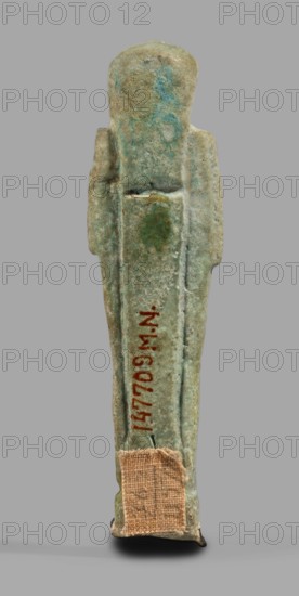Ushabti, Wigs, funerary figurine, mummy-shaped standing on a base, back supported by a pillar/pilaster. Arms crossed on the chest, right folded over left, both hands holding hoes. On the back is a bag slung over the left shoulder on a single string. Head with a three-part smooth wig back strand longer than front strands. Long, straight, wide beard. Large eyes, wide nose, mouth set in a grimace, very large ears. No inscription. Late Period, 664–332 BC, Egypt. Shabti; height 7.7 cm, width 2.1 cm., A narrow upright figurine seen from the back against a plain gray background, appearing weathered with a rough surface; the object shows a range of colors including greenish-blue, turquoise, pale green, gray, beige, and small areas of brown and reddish tones; the shape has a rounded head, straight shoulders, and long straight sides with slight raised ridges running vertically; there is a rectangular patch of coarse beige fabric adhered near the lower end and a small darkened area at the base; t...
