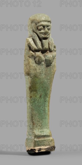 Ushabti, Mummy-shaped funerary figurine standing on a base, back supported by a pillar/pilaster. Arms crossed on the chest, the right folded over the left; both hands holding hoes. A bag is on the back, slung over the left shoulder on a single string. Wearing a three-part smooth wig with the back strand longer than the front strands. Long, straight, wide beard; large eyes, wide nose, mouth set in a grimace, very large ears. No inscription. Late Period, Egypt, 664–332 BC. Shabti; height 7.7 cm, width 2.1 cm., A small statuette of a standing figure with a long body and a stylized head and shoulders, shown against a plain gray background; the figure appears to wear a headdress with a fringe and has a defined face with a pronounced chin and brow, and arms crossed over the chest; the surface shows a weathered, mottled texture with visible pitting and flaking; colors visible include shades of green, turquoise, pale blue, beige, brown, and touches of darker brown or black at the base; the bas...