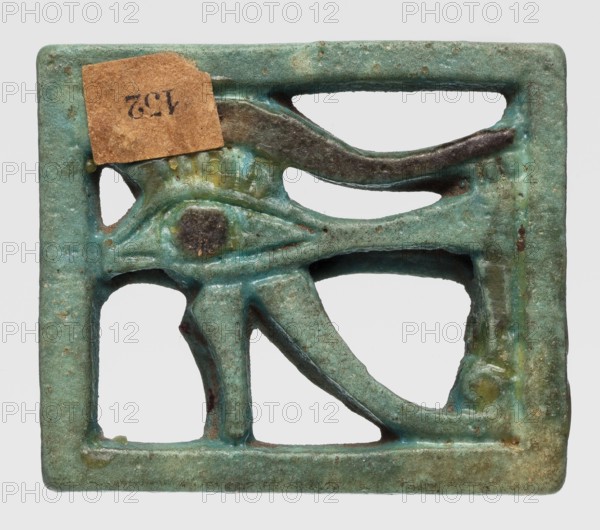 Amulet - Eye of Horus, Openwork Eye of Horus in a rectangular frame. Iris and eyebrow black, the rest green-blue. ca. 945–656 BC; Third Intermediate Period, 22nd–25th Dynasty, found in Edfu; amulet, height 2.9 cm, width 3.4 cm., A small rectangular object with open cutouts forming a stylized eye motif, rendered in a weathered bluish-green with areas of darker green and brown patina; a rough square of tan paper adhered near the top left; the surface shows small pits and spots of darker brown and black, with some lighter green and yellowish discoloration around raised details.