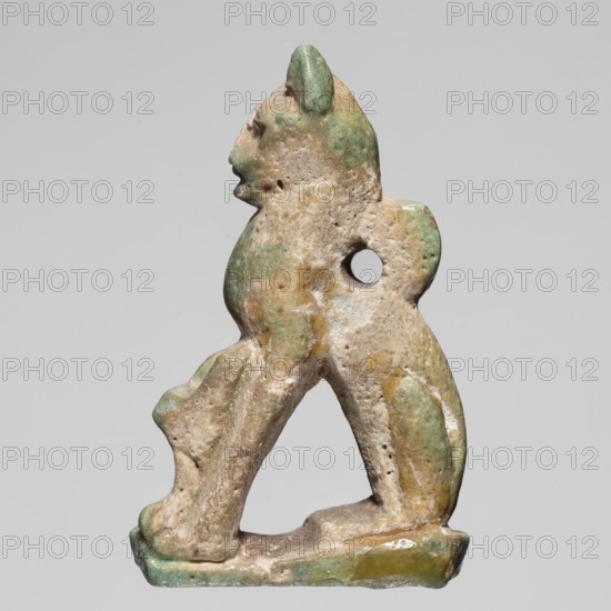 Amulet — cat; maker, cat-shaped amulet; pendant; Egyptian, ca. 945–525 BC 22nd–26th Dynasty; materials not specified; height 3.3 cm, width 2 cm., A small sculptural object depicting an animal-like figure in a seated or crouching pose, with a rounded head, pronounced snout, and upright ears; the form has a hole through the torso area and sits on a flat base. The surface shows uneven texture with weathered and pitted areas, and visible coloration in shades of green, beige, tan, and hints of yellow, with some areas appearing smoother and glossier while others look rough and matte. The background is a plain light gray.