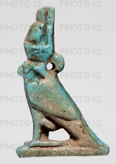 Amulet - Horus; falcons, birds, sacred animals, amulets, Horus myth.. Amulet of Horus in the form of a falcon sitting on a rectangular base with a double crown on its head. 664–332 BC; Late Period, Egypt. Amulet; height 4.9 cm, width 2.5 cm., A small sculpted object shaped like a perched bird with a hole through its head, rendered in turquoise and pale blue glaze with areas of brown and beige patina and wear, showing rough textured spots and smoother glossy patches, with the bird sitting on a flat base and a hollowed area beneath its body, photographed against a light gray background.