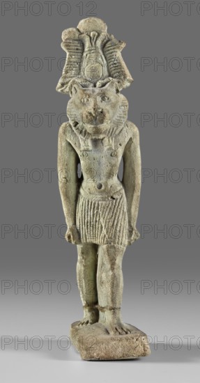 Amulet - god Mahes. Figurine of a striding lion-headed male deity wearing an atef crown, dressed in a pleated loincloth; pillars on the back. Egyptian deity Mahes mythology. 1st century; Roman period, Egypt. Amulet, height 7.4 cm, width 2.2 cm., A standing figurine of a human body with a feline head wearing a tall headdress, depicted in a frontal pose with arms down and hands beside the thighs, dressed in a short pleated skirt and wearing a collar around the neck, mounted on a rectangular base. The surface shows a weathered, textured appearance with areas of erosion and small chips. Visible colors include pale green, beige, light brown, and hints of gray, with darker shadowed tones in recessed areas. The background is a uniform medium gray.