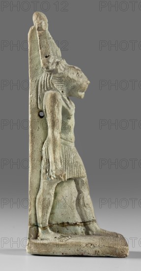 Amulet - god Mahes; figurine of a striding lion-headed male deity wearing an atef crown, dressed in a pleated loincloth; pillars on the back; amulet; Mahes mythology; 1st century, Roman period, Egypt; height 7.4 cm, width 2.2 cm., A weathered carved figure in profile standing on a rectangular base, showing a humanoid body with a feline head and an upright back pillar; the figure wears a pleated skirt and has a defined arm hanging alongside the torso, with visible grooves suggesting hair or a headdress; surface texture is pitted and rough with areas of erosion and small holes; colors include pale greenish-blue, beige, light gray, and muted brown tones, with darker gray used for the background.