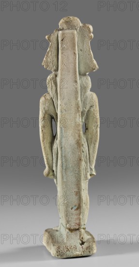 Amulet - god Mahes; Mahes mythology. Figurine of a striding lion-headed male deity wearing an atef crown, dressed in a pleated loincloth. Pillars on the back. 1st century; Roman period, Egypt. Amulet; height 7.4 cm, width 2.2 cm., A weathered standing figure seen from the back, rendered in a pale greenish-beige material with areas of light brown and bluish-green discoloration; the figure has a long vertical central support running from head to base, a flared headdress, rounded shoulders, and stylized arms hanging at the sides with hands near the hips; the surface appears rough and pitted with small chips and worn edges, and the figure stands on a rectangular base that shares the same muted pale green, beige, and brown tones against a neutral gray background.
