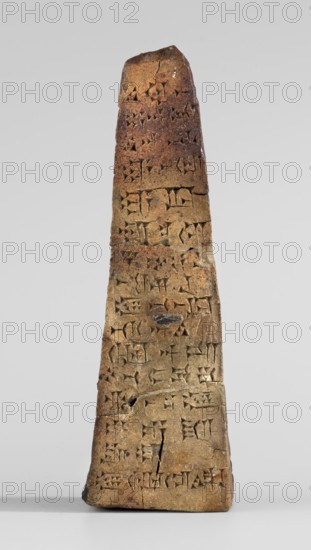Votive obelisk with an inscription of King Ashurbanipal forgery with a copy of an ancient text, cuneiform writing, Ashurbanipal king of Assyria; ca. 668–ca. 631 BC, votive offering, obelisk, inscription, forgery. Clay votive obelisk tapering toward the top; cuneiform text inscribed on all four faces. Dating of copied inscription content: c. 668–c. 631 BC Neo-Assyrian period; object itself dated to 2nd half 19th century–1st half 20th century forgery. Inscription based on texts from Iraq Quyunjik, Nineveh. Document/dedication. Height 13.1 cm; width 3.7 cm; thickness 4.2 cm., A tall, narrow stone object with a tapered, roughly triangular silhouette standing upright on a flat surface, showing a textured, weathered surface with carved indentations across its face; the stone displays a range of colors including brown, tan, beige, and darker brown patches, with some small worn or chipped areas and visible hairline cracks, while the background is a smooth light gray.