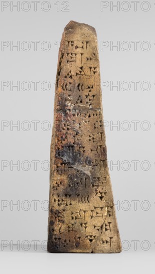 Votive obelisk with an inscription of King Ashurbanipal forgery with a copy of an ancient text, cuneiform writing, Ashurbanipal king of Assyria; ca. 668–ca. 631 BC, votive offering obelisk. Clay obelisk tapering toward the top with cuneiform text inscribed on all four faces. Dating of copied inscriptions: c. 668–c. 631 BC Neo-Assyrian period; object itself dated to 2nd half 19th century–1st half 20th century forgery. Inscription based on texts from Iraq Quyunjik, Nineveh. Function: dedication/document. Dimensions: height 13.1 cm; width 3.7 cm; thickness 4.2 cm., A tapered, upright stone object with a rough, weathered surface stands against a neutral light gray background; the object is primarily tan and brown with patches of darker brown and black discoloration and small areas of reddish coloration, and its surface shows numerous small triangular and wedge-shaped indentations arranged across the face; the top is irregular and slightly angled, the sides slope inward toward the top, and ...
