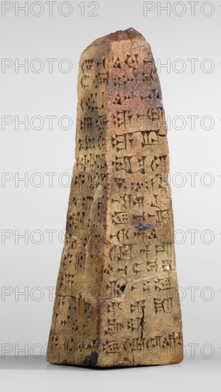 Votive obelisk with an inscription of King Ashurbanipal forgery with a copy of an ancient text, cuneiform writing, Ashurbanipal king of Assyria; ca. 668–ca. 631 BC, votive offerings, obelisks, inscriptions, forgeries. Clay votive obelisk of King Ashurbanipal. The obelisk tapers towards the top. Cuneiform text inscribed on all four faces of the object. Dating of copied inscriptions: c. 668–c. 631 BC Neo-Assyrian period; forgery dated to 2nd half 19th century–1st half 20th century. Inscription based on texts from Iraq Quyunjik, Nineveh. Forgery probably from the Middle East. Document; dedication. Height 13.1 cm, width 3.7 cm, thickness 4.2 cm., A tall, tapered stone object with an uneven, pyramidal shape stands upright against a plain light gray background, showing a rough, weathered surface with visible carved impressions across its faces. The stone displays a range of earthy colors including beige, tan, brown, and rusty orange, with some darker brown and charcoal spots and patches. The...