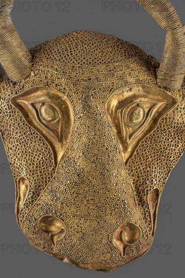 Protoma; bull's head relief mask made in gold using repoussé, granulation, and filigree. Central nostril area features granulation and filigree; cheeks and top use filigree only. Eyes, nostrils, and muzzle are repoussé and bordered with filigree. Small ears and horns are plastically modeled horns soldered on. Reverse closed with an undecorated gold sheet bearing an irregular hole removal of a fastening element. Date: ca. 1450–ca. 1100 BC Middle Elamite period, — alternative datings proposed to the Achaemenid period 600–539 BC and to the second half of the 2nd millennium BC. Place of origin: Elam, Iran. Decoration; height 9.5 cm, width 7.8 cm, depth 2.5 cm; weight 48 g., A close-up of a metallic decorative object shaped like an animal face, showing textured surfaces and raised features in gold tones against a gray background; the surface is covered in a dense pattern of small circular bumps and rings, with smoother, sculpted areas forming stylized eyes, nostrils, and other facial contou...