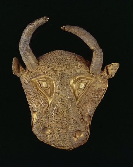 Protoma bull head relief mask. Gold; repoussé, granulation, filigree, soldering. Central nostril area with granulation and filigree; cheeks and top with filigree; eyes, nostrils and muzzle repoussé and bordered with filigree. Small ears and plastically modeled horns horns soldered on. Reverse closed with an undecorated gold sheet with an irregular hole in two-thirds of its height from removal of a fastening element. Ca. 1450–1100 BC Middle Elamite period ?; alternative proposed datings include Achaemenid period 600–539 BC and second half of the 2nd millennium BC. Origin: Elam/Iran. Decoration. Height 9.5 cm; width 7.8 cm; depth 2.5 cm; weight 48 g., A golden decorative mask in the shape of an animal head set against a black background, featuring a textured surface made of many small circular patterns and smooth raised areas, with a pair of curved horns wrapped in a fine coil-like texture, prominent sculpted eye shapes and a pronounced snout, and visible colors including gold tones and ...