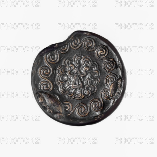Stamp seal, unknown ancient Hittite workshop. Hittite stamp seal made of hematite. Carefully crafted handle with eight surfaces tapering upward, terminating in a ring and a spherical grip with a horizontally drilled hole. Circular base bears two carved ornament circles: a larger spiral tendril and a smaller inner braid. Two characters imitating Hittite hieroglyphic writing were added to the smaller circle in the 19th century. Date: 17th–14th century BC; Old Hittite period Hittite hieroglyphic characters added in the 19th century. Provenance: Karahöyük Adiyaman Province, Turkey; Hittite Empire c.1650–c.1178 BC. Medium: hematite. Type: seal. Dimensions: height 3.1 cm; width 3.3 cm., A round, dark object with a glossy black surface and worn bronze highlights shows a raised central floral or rosette motif surrounded by a ring of repeating spiral or wave patterns; the edges are irregular with a few chipped or dented areas exposing lighter bronze tones, and the overall palette includes black...