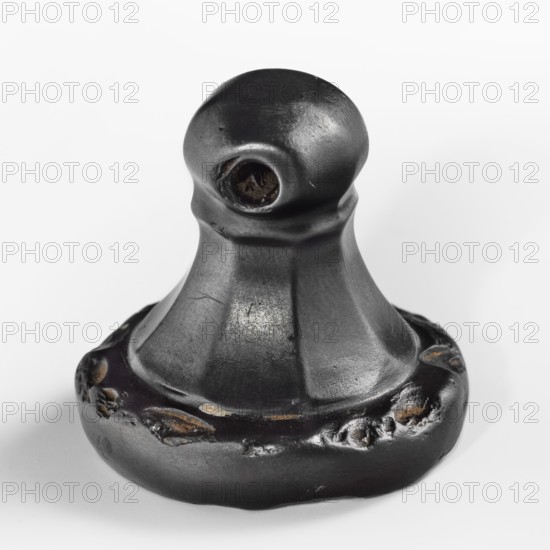 Hittite stamp seal made of hematite. Carefully crafted handle with eight tapering faces ending in a ring and a spherical grip with a horizontal perforation. Circular base carved with two ornamented concentric circles: a larger spiral-tendril motif and a smaller braided motif. Two characters imitating Hittite hieroglyphic writing were added in the 19th century. Old Hittite period, 17th–14th century BC. From Karahöyük Adiyaman Province, Turkey, Hittite Empire. Seal; height 3.1 cm, width 3.3 cm., A dark metallic object with a rounded top and a flared base sits against a white background; the surface shows a matte black finish with areas of darker and lighter gray sheen, rough patches, and small worn spots revealing brownish tones, and there is a circular recess near the top with a textured dark interior.