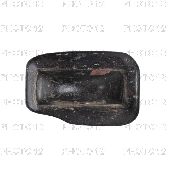 Stamp seal with a depiction of a bull's head. Eastern Cypriot, stamp seal with a full, arched handle with a horizontally drilled hole. On its rectangular base is carved a schematic bull's head with small ears and long, curved horns, between which is a jug with a triangular belly and a long, narrow neck with a handle. On either side of the bull's muzzle are carved short arrows with triangular heads pointing downwards—possibly a simplified representation of snakes. The whole is framed with beadwork. Cypriot glyptics; bull heads iconography. 14th–13th century BC, Late Bronze Age c. 1450–1250 BC. Cyprus. Seal. Height 3.2 cm, width 5.4 cm, depth 3.5 cm., A worn dark object with an irregular rectangular shape and rounded corners, showing a smooth, slightly glossy surface with patches of lighter abrasion and scuff marks; the object has a recessed central area forming a shallow cavity and a raised rim around it, with small areas of brownish discoloration and faint hints of lighter gray where t...