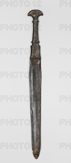 Dagger, unknown Luristan workshop. Dagger with a cast blade and a mushroom-shaped hilt with a rimmed mount; the raised edges of the mount served to hold a lost hilt casing, possibly of bone, wood, horn, or stone. The hilt shaft is richly profiled and flows directly into the blade with a centric point. Behind the short, rectangular base, blunted on both sides, the double-edged blade tapers evenly toward the tip. A slightly convex ridge runs through the center of the blade and splits along one-third of its length toward the base, creating a triangular flat field. 9th–6th century BC; Iron Period II 1st half of the 1st millennium BC. Luristan, Iran. Weapon. Height 33.8 cm; width 2.9 cm; depth 0.8 cm; weight 194 g., A long vertical metallic object with an elongated straight blade and a distinct handle at the top, displayed against a light gray background. The handle has a rounded top and a narrow neck with sculpted inward curves, showing an aged, slightly irregular surface. The blade is str...