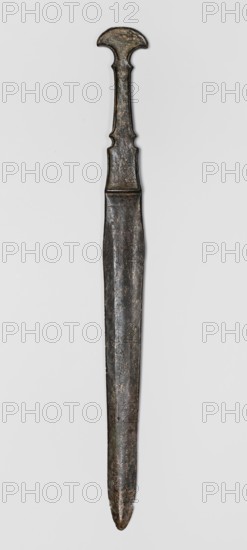 Dagger, unknown Luristan workshop. Dagger with a cast blade and a mushroom-shaped hilt with a rimmed mount; the raised edges of the mount served to hold a lost hilt casing possibly bone, wood, horn, or stone. The hilt shaft is richly profiled and flows directly into the blade with a centric point. Behind the short, rectangular base, blunted on both sides, the double-edged blade tapers evenly towards the tip. A slightly convex ridge runs through the center of the blade, which splits along one-third of its length towards the base, creating a triangular flat field. 9th–6th century BC; Iron Period II 1st half of the 1st millennium BC. Luristan, Iran. Weapon. Height 33.8 cm; width 2.9 cm; depth 0.8 cm; weight 194 g., A single elongated metal object with a narrow pointed end and a wider decorative top, shown against a light background; the object has a long straight shaft that tapers slightly toward the tip and displays an uneven, textured surface with signs of wear; the upper portion featur...