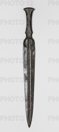 Dagger, unknown Luristan workshop. A dagger with a blade cast together with a mushroom-shaped hilt, with a rimmed mount whose raised edges served to hold a hilt casing that has not been preserved. The casing may have been made of bone, wood, horn, or stone. In the central part of the mount, short arched sheet-metal protrusions are bent inward to secure the casing. The hilt merges directly into a lanceolate, double-edged blade with a central point. The base of the blade is in the form of a convex ring, open at the bottom. A convex ridge with narrow grooves on the sides runs through the center of the blade from tip to base. 9th–6th century BC; Iron Period II end of the 2nd millennium–first half of the 1st millennium BC. Luristan, Iran. Weapon. Height 28.5 cm; width 2.9 cm; thickness 1.1 cm; weight 116 g., A long metallic object with a dark brownish-black patina, showing areas of lighter brown and muted gray where the surface is worn, is centered against a light gray background; it tapers...