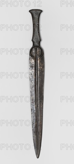 Dagger, unknown Luristan workshop. A dagger with a blade cast together with a mushroom-shaped hilt, with a rimmed mount whose raised edges held a now-lost hilt casing possibly bone, wood, horn, or stone. In the central part of the mount, short arched sheet-metal protrusions are bent inward to secure the casing. The hilt merges directly into a lanceolate, double-edged blade with a central point. The base of the blade is a convex ring open at the bottom. A convex ridge with narrow lateral grooves runs along the blade from tip to base. 9th–6th century BC; Iron Period II end of the 2nd millennium–first half of the 1st millennium BC. Luristan, Iran. Weapon. Height 28.5 cm; width 2.9 cm; thickness 1.1 cm; weight 116 g., A long metallic object is shown vertically against a pale background, with a dark gray to black surface and areas of lighter gray and brownish patches; the object tapers to a pointed tip and has a symmetrical, flattened blade with a central raised ridge; near the top a handle...