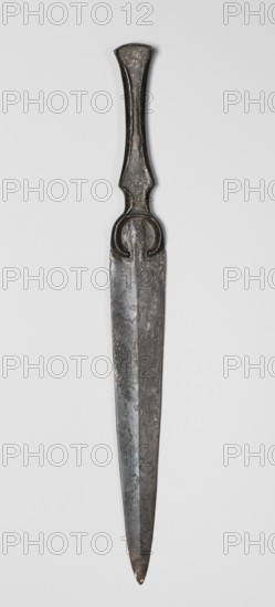 Dagger, unknown Luristan workshop. Short dagger with a blade cast together with a mushroom-shaped hilt and a rimmed mount whose raised edges held a now-lost hilt casing possibly bone, wood, horn, or stone. In the central part of the mount, short arched sheet-metal protrusions are bent inward to secure the casing. The hilt merges directly into a lanceolate, double-edged blade with a central point. The base of the blade is a convex ring open at the bottom. A prominent ridge line runs through the center of the blade from tip to base, widening in the ring area to create a small convex triangular zone. 9th–6th century BC; Iron Age II 1st half of the 1st millennium BC. Luristan, Iran. Weapon. Height 24 cm; width 2.5 cm; depth 1.1 cm; weight 76 g., A long metal object with a narrow pointed blade and an elongated handle, shown vertically against a plain light gray background; the object appears dark gray with areas of lighter gray and subtle greenish and brownish patina, with a rounded tip on ...
