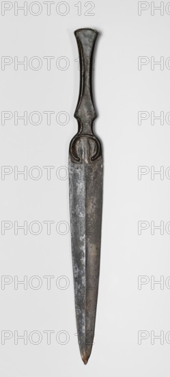 Dagger, unknown Luristan workshop. Short dagger with a blade cast together with a mushroom-shaped hilt and a rimmed mount; the raised edges of the mount served to hold a hilt casing that has not been preserved possibly bone, wood, horn, or stone. In the central part of the mount, short arched sheet-metal protrusions are bent inward to secure the casing. The hilt merges directly into a lanceolate, double-edged blade with a central point. The base of the blade is a convex ring, open at the bottom. A prominent ridge runs through the center of the blade from the tip to the base, widening in the ring area to create a small convex triangular region. Luristan bronzes; daggers, edged weapons; Iranian art; 9th–6th century BC Iron Period II, 1st half of the 1st millennium BC. Luristan, Iran. Weapon. Height 24 cm; width 2.5 cm; depth 1.1 cm; weight 76 g., A single metal dagger is shown vertically against a light gray background, with a dark, slightly flared handle that tapers into a narrow neck a...