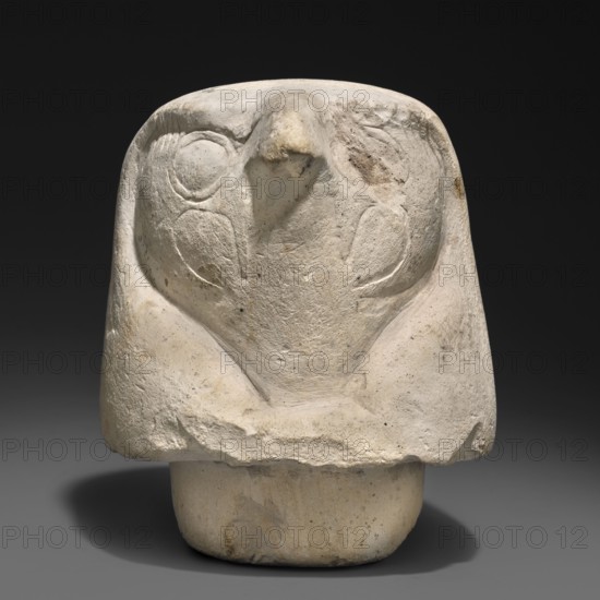 Canopic jar lid — falcon head;, Canopic jar lid in the shape of a plastically modeled falcon head, with details around the beak and eyes rendered with delicate modeling; underside with a wide tenon securing the lid in the urn's opening. Sons of Horus mythology; Kebehsenuf mythology; falcon iconography; Egyptian art; Egyptian sculpture. Late Period, Egypt, 664–332 BC. Canopic jar; height 11 cm, width 9 cm., A carved pale beige stone object sits against a dark gray to black background, showing a rounded top and a stylized front with carved circular shallow depressions and a central protruding ridge. The surface shows rough, pitted texture with areas of smoother wear and small darker gray and brown spots. The base is cylindrical and slightly lighter in color with similar speckling, and the object casts a diffuse shadow on the neutral gray surface below.