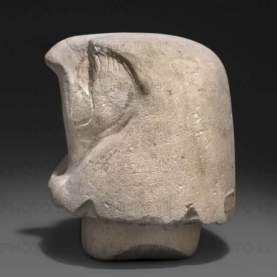 Canopic jar lid — falcon head; canopic jars; Sons of Horus mythology; falcon iconography; Egyptian art; Egyptian sculpture; Kebehsenuf mythology. Canopic jar lid in the shape of a plastically modeled falcon head, with details around the beak and eyes rendered with delicate modeling. On the underside, a wide tenon securing the lid in the urn's opening. 664–332 BC, Late Period, Egypt. Canopic jar; height 11 cm, width 9 cm., A pale beige stone sculpture shown from a side angle on a dark gray background, with a rounded top and a carved concave area suggesting a stylized face profile; the surface is textured with scratches and small pits, and there are smoother worn areas and a few rough chips along the lower edge. The base is slightly darker beige and sits in a soft shadow; overall colors visible include off-white, beige, light gray, dark gray, and a hint of brown in worn spots.