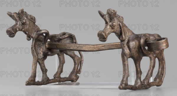 Bit, unknown Luristan workshop. The bit consists of a rigid bar and two cheek plates depicting a horse in profile. The bar, with a quadrangular cross-section, is slightly curved and has chamfered edges. Its ends are forged into a ribbon, which is wrapped in oppositely facing rings. The bar is inserted into two round holes in the cheek plates, which are located in the animals' torsos; externally they form circular rings protruding from the background, occupying the entire surface of the plates in this location. The cheek plates are modeled on one side; only the animal heads and necks are modeled on both sides. Maker attribution: Antoni Mierzejewski . Date: 9th–6th century BC; Iron Period II c. 1000–601 BC. Country/place of find and manufacture: Luristan, Iran. Function: element of a horse harness. Dimensions: height 8.6 cm; width 20 cm; depth 13.8 cm; weight 480 g., A bronze-colored sculptural object depicting a pair of stylized animals facing opposite directions and connected by a hori...