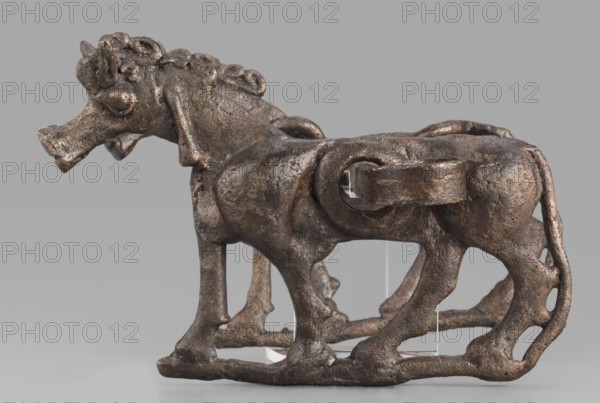 Bit, unknown Luristan workshop. The bit consists of a rigid bar and two cheek plates depicting a horse in profile. The bar, with a quadrangular cross-section, is slightly curved and has chamfered edges. Its ends are forged into a ribbon, which is wrapped in oppositely facing rings. The bar is inserted into two round holes in the cheek plates, located in the animals' torsos; externally they form circular rings protruding from the background, occupying the entire surface of the plates at that location. The cheek plates are modeled on one side; only the animal heads and necks are modeled on both sides. Attributed to Antoni Mierzejewski. Dated 9th–6th century BC; Iron Period II c. 1000–601 BC. Find place: Luristan, Iran; likely made in Luristan. Function: element of a horse harness. Materials: Luristan bronzes. Dimensions: height 8.6 cm, width 20 cm, depth 13.8 cm; weight 480 g., A small sculpted object in shades of brown and dark bronze appears against a neutral gray background; it depict...