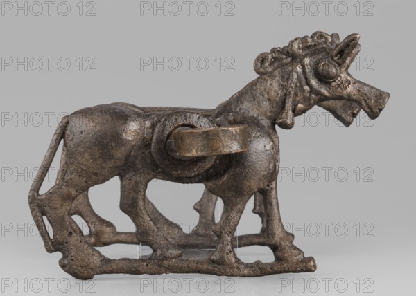 Bit, unknown Luristan workshop. The bit consists of a rigid bar and two cheek plates depicting a horse in profile. The bar, with a quadrangular cross-section, is slightly curved and has chamfered edges. Its ends are forged into a ribbon, which is wrapped in oppositely facing rings. The bar is inserted into two round holes in the cheek plates, which are located in the animals' torsos. Externally, they form circular rings protruding from the background, occupying the entire surface of the plates in this location. The cheek plates are modeled on one side; only the animal heads and necks are modeled on both sides. Attributed to Antoni Mierzejewski. 9th–6th century BC; Iron Period II c. 1000–601 BC. Iran, Luristan place of find and manufacture. Element of a horse harness. Height 8.6 cm; width 20 cm; depth 13.8 cm; weight 480 g., A small sculpted animal figure shown in profile on a flat surface, cast in a dark brown metallic material with a slightly rough, pitted texture and areas of lighter...
