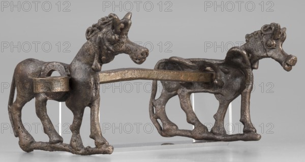 Bit, unknown Luristan workshop. The bit consists of a rigid bar and two cheek plates depicting a horse in profile. The bar, with a quadrangular cross-section, is slightly curved and has chamfered edges. Its ends are forged into a ribbon, which is wrapped in oppositely facing rings. The bar is inserted into two round holes in the cheek plates, located in the animals' torsos; externally they form circular rings protruding from the background, occupying the entire surface of the plates in this location. The cheek plates are modeled on one side; only the animal heads and necks are modeled on both sides. Attributed to Antoni Mierzejewski, 9th–6th century BC; Iron Period II c. 1000–601 BC. Iran, Luristan find/place of manufacture. Element of a horse harness. Height 8.6 cm; width 20 cm; depth 13.8 cm; weight 480 g., A sculptural object showing a pair of animal figures joined by a horizontal bar, each figure standing on a connected base. The figures have elongated necks, rounded muzzles, promi...