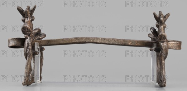Bit, unknown Luristan workshop. The bit consists of a rigid bar and two cheek plates depicting a horse in profile. The bar, with a quadrangular cross-section, is slightly curved and has chamfered edges; its ends are forged into a ribbon wrapped in oppositely facing rings. The bar is inserted into two round holes in the cheek plates located in the animals' torsos; externally these form circular rings protruding from the background, occupying the entire surface of the plates at that location. The cheek plates are modeled on one side; only the animal heads and necks are modeled on both sides. Attributed to Antoni Mierzejewski, 9th–6th century BC; Iron Period II c. 1000–601 BC. Findplace/manufacture: Luristan, Iran. Element of a horse harness. Height 8.6 cm; width 20 cm; depth 13.8 cm; weight 480 g., A single metal horizontal bar with a textured, aged surface rests on a clear plastic support, flanked at each end by sculpted vertical elements that resemble stylized animal heads with elongat...