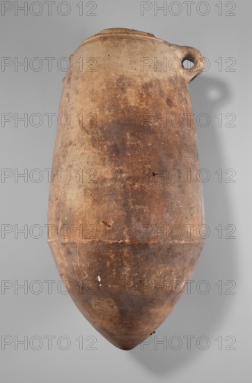 Amphora, Amphora with a cylindrical body slightly tapering toward the top. Pointed bottom, annular spout, two small handles one damaged. Egyptian ceramics, excavated in Edfu; vessel; height 55 cm, width 26.7 cm., A large, elongated clay vessel with a rounded, pointed base and a narrow, slightly flared rim, showing a small loop handle near the top; the surface is textured and worn with visible patches of roughness and small chips, and the colors visible include shades of brown, tan, beige, and darker brown spots, set against a neutral light gray background.