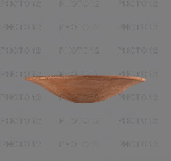 Bowl. Low bowl with irregular, strongly flaring walls and a rounded bottom. Rounded rim, slightly rolled outward. Surface smoothed and covered with a layer of red engobe. Bottom worked with a tool. Hand-made. Vessel glued together from several fragments. Comparative material: similar forms dated c. 2686–2181 BC, Old Kingdom, Edfu; vessel dimensions: height 6.1 cm, diameter 28.3 cm rim and body. Maker: unknown., A shallow, round, terracotta-colored bowl with a wide, slightly flared rim is shown against a flat gray background; the bowl's surface displays a few small pits and subtle variations in reddish-brown and muted orange tones, with some darker speckles and lighter worn areas, and the interior and exterior share the same earthy coloration and rough, handmade texture.