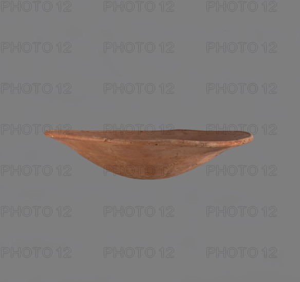 Bowl. Low bowl with irregular, strongly flaring walls and a rounded bottom. Rounded rim, slightly rolled outward. Surface smoothed and covered with a layer of red engobe. Bottom worked with a tool. Hand-made. Vessel glued together from several fragments. Egyptian pottery, clay vessel; ceramics, bowl. Comparative typology: Reisner, Mycerinus, type XXVII–XXVIII, fig. 72 2–5; c. 2686–2181 BC Old Kingdom, Edfu. Dimensions: height 6.1 cm; diameter rim 28.3 cm; diameter body 28.3 cm., A shallow, rounded clay bowl with an irregular rim is centered against a plain gray background; the bowl has a terracotta color with variations of reddish-brown and muted orange, showing a slightly rough texture and small surface imperfections.