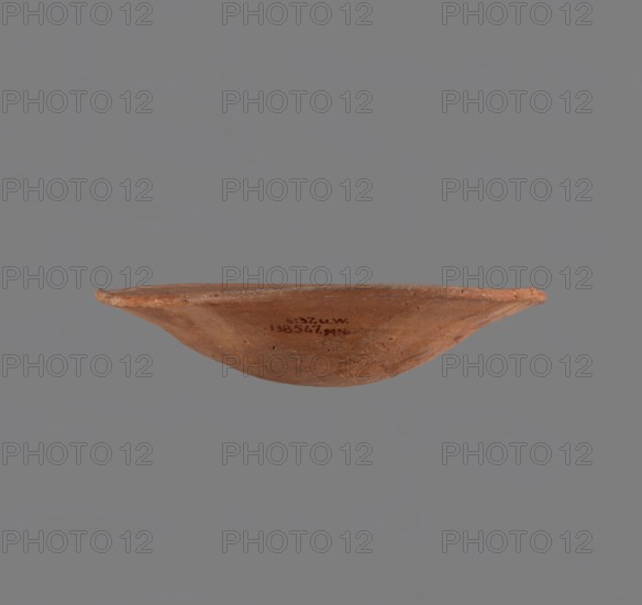 Bowl. Low bowl with irregular, strongly flaring walls and a rounded bottom. Rounded rim, slightly rolled outward. Surface smoothed and covered with a layer of red engobe. Bottom worked with a tool. Hand-made. Vessel glued together from several fragments. Egyptian pottery, clay vessel, ceramic bowl. Comparative material: Reisner George A., Mycerinus... type XXVII and XXVIII, fig. 72 2-5, c. 2686–c. 2181 BC; Old Kingdom, Edfu; vessel height 6.1 cm, rim diameter 28.3 cm, body diameter 28.3 cm. Creator/classification unknown., A shallow, rounded bowl photographed from the side against a gray background, showing an earthy terracotta surface with a few darker spots and irregularities; the rim is slightly uneven and the visible colors include reddish-brown, terracotta orange, and muted gray.