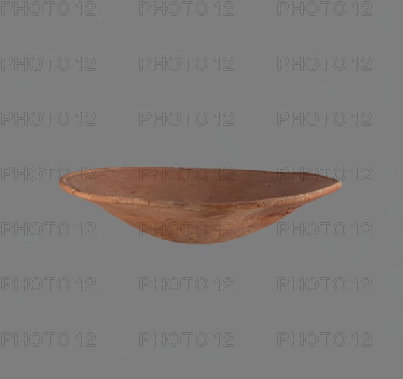 Bowl. Low bowl with irregular, strongly flaring walls and a rounded bottom. Rounded rim, slightly rolled outward. Surface smoothed and covered with a layer of red engobe. Bottom worked with a tool. Hand-made. Vessel glued together from several fragments. Egyptian pottery, clay/ceramic vessel, bowl. Old Kingdom, c. 2686–2181 BC. Height 6.1 cm; diameter rim/body 28.3 cm., Edfu place of origin., A shallow, round earthenware bowl with a slightly uneven rim and a smooth interior surface, shown against a plain gray background; the bowl appears in shades of reddish-brown and terra cotta with some darker speckles and subtle variations in tone, and the background is medium gray.