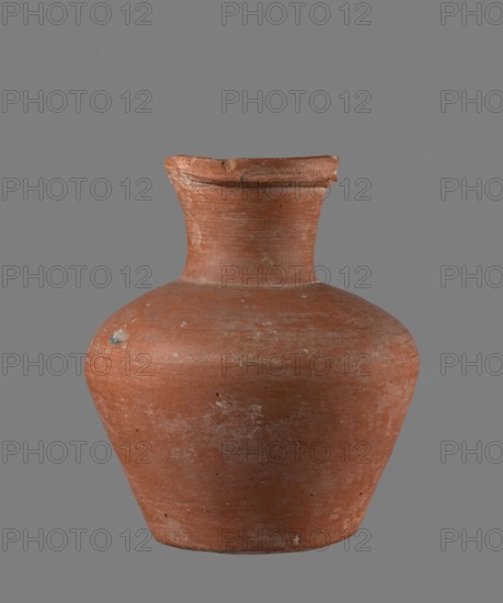 Jug with biconical belly, widest at mid-height; broad, gently sloping shoulders; wide neck slightly widening toward a profiled, irregular spout that is slightly raised and extended on one side forming a small secondary spout. Spout nearly triangular in cross-section with a ~1 mm indentation at the center of the outer profile. Bottom very slightly rounded. Height of neck about one-third of total vessel height. Made of Nile clay NB1? — Vienna System. Neck formed separately and joined; join traces erased. Outer surface smoothed, covered with engobe and polished; inner surface smoothed except for the neck, which is also engobed and polished. Reddish-brown color. Good preservation; rim slightly chipped in two places and engobe worn off in places on the outer surface. Local workshop: Edfu. Comparative typology: parallels to Old Kingdom types ca. 2686–2181 BCE., A worn terracotta vessel with a rounded body that tapers toward a narrower base and a short, slightly flared neck; the surface shows...