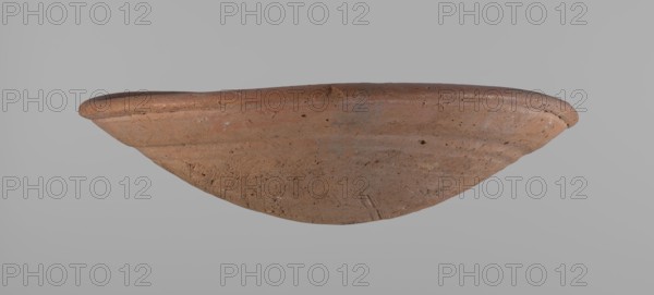 Shallow bowl of rounded, asymmetrical shape with rounded, flaring, irregular rim. Surface level; lower part finished to approximately half its height with a knife or similar tool. Slightly above the middle, a horizontal line likely from placement on a stand. Dark red clay; complete. From Edfu — local workshop. Comparative material: A. Reisner, A History of the City of the Giza Necropolis I, fig. 227, 35-8-16, ca. 2686–2181 BC Old Kingdom. Dimensions: height 7.2 cm, diameter 26.2 cm rim., A shallow, rounded ceramic bowl is shown from a side angle against a plain gray background; the bowl has an earthy brown color with variations of lighter and darker brown tones and small darker flecks, a slightly raised rim with a smoother texture, and a surface marked by subtle scratches, pits, and unevenness indicative of its material.